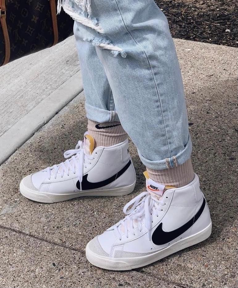 d4432fb2-3942-4e7c-bdf1-afb60738fd7f_00158c04-6f0a-4f31-905e-8964c07ec286 | UAE SPORT STORE Person wearing white Nike high-top sneakers with a black swoosh, beige socks, and light-wash rolled jeans on a sidewalk.