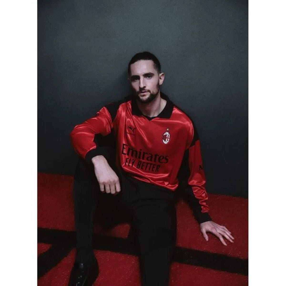 Man wearing a red AC Milan football jersey sits against a dark backdrop, looking at the camera with a neutral expression.
