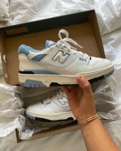 d471aca0-44db-4de2-857a-4a07d7dc0b4d_5dab3304-65bc-4b09-b44f-dc1ee0f5c523 | UAE SPORT STORE White and light blue New Balance 550 sneaker being held over its shoe box, showing the side with the large 'N' logo and perforated panels.