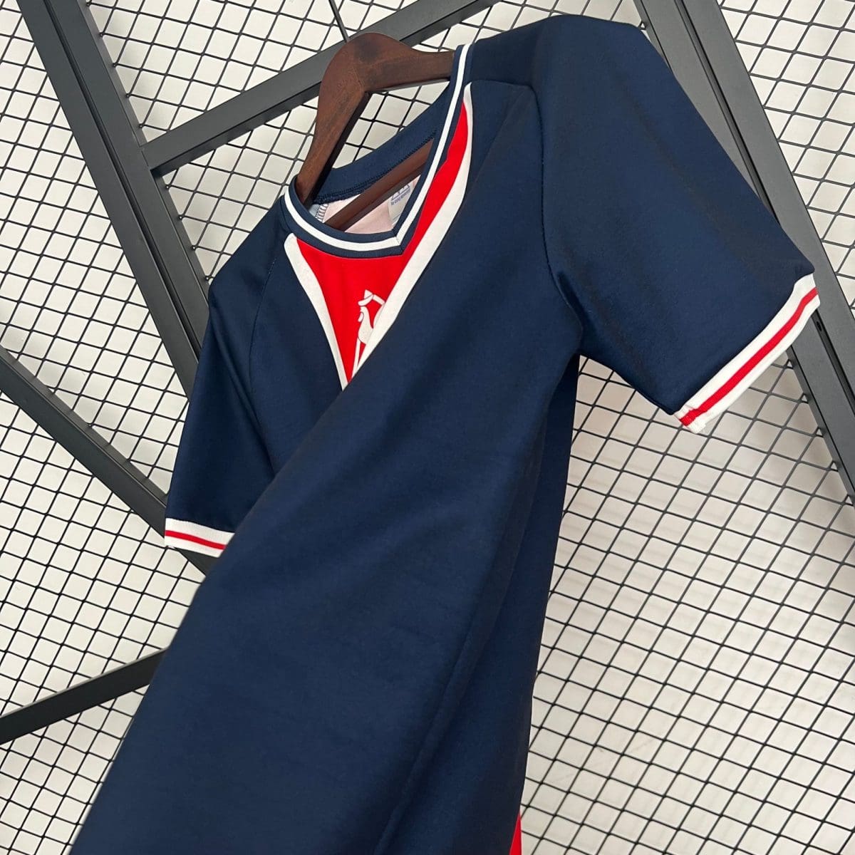 Navy blue varsity-style jacket on a wooden hanger, with white and red trim, displayed against a metal grid backdrop in a store setting.