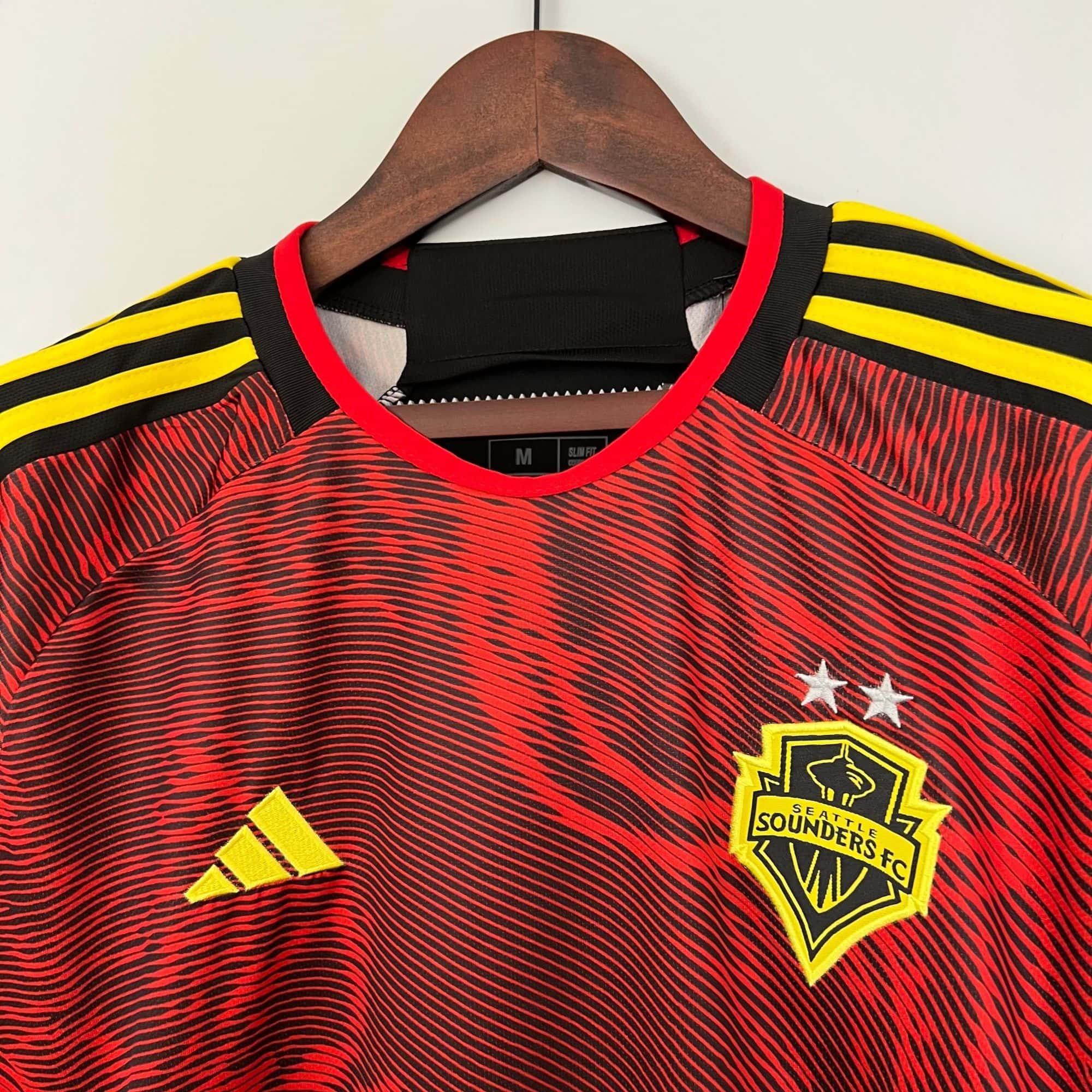 d4ce7178_3d77fe9f-1b71-49d7-ae27-4dcd002b444d | UAE SPORT STORE Red and black striped soccer jersey on a wooden hanger, with yellow shoulder stripes and a Seattle Sounders FC crest on the chest.