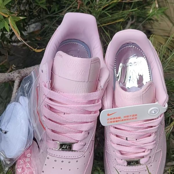 Two light-pink Nike Air Force 1 sneakers with pink laces on grass, showing perforated toes and a white tag on the right shoe.