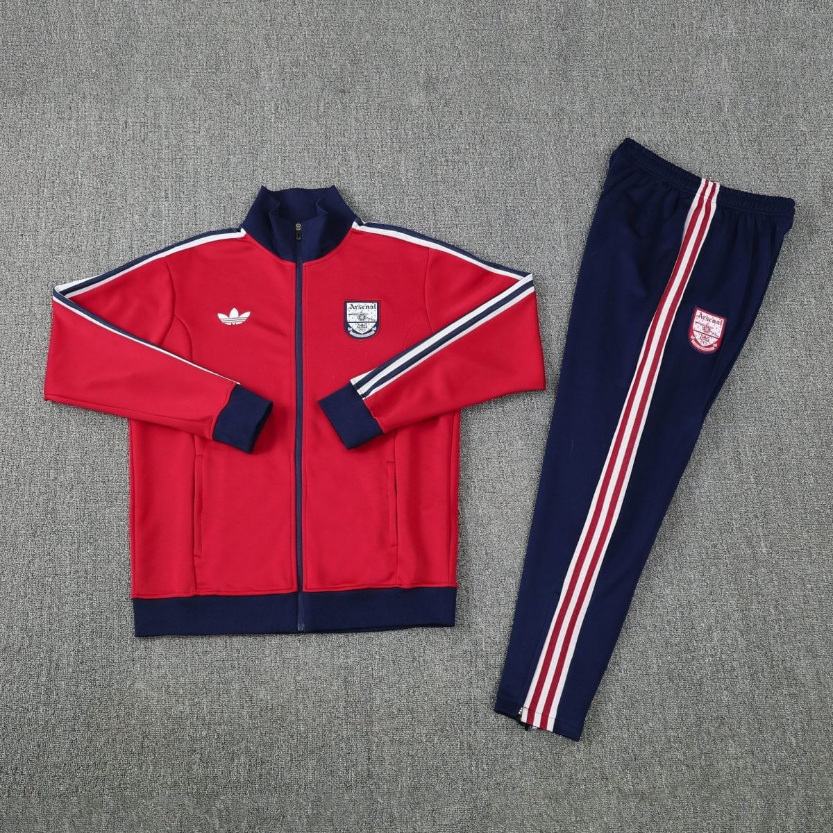 Red Arsenal track jacket with navy cuffs and white stripes laid beside matching navy track pants on a gray carpet, outfit ready to wear.