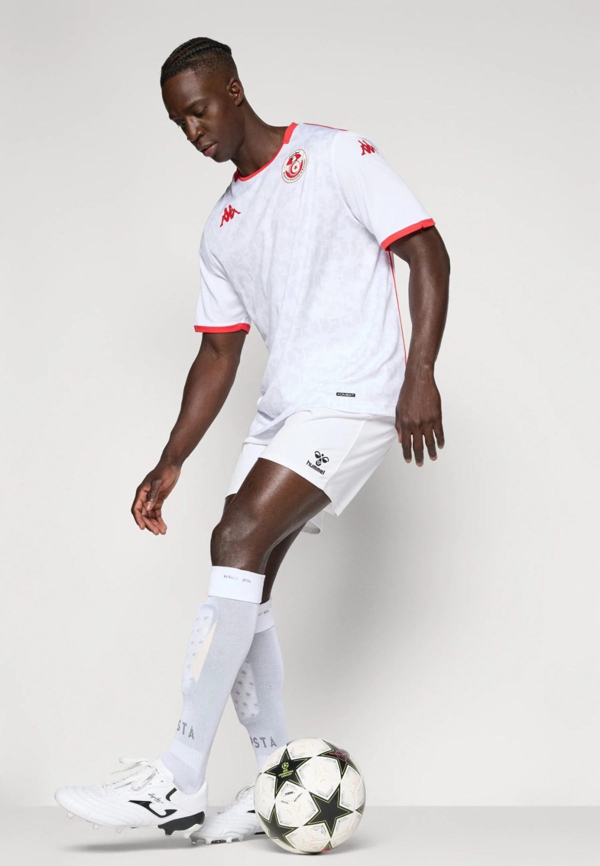 Male soccer player in a white jersey with red trim and crest, about to kick a patterned soccer ball against a neutral backdrop.