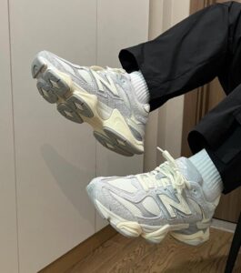 Light gray New Balance sneakers with a chunky off-white sole, worn with white socks, indoors on a wooden floor