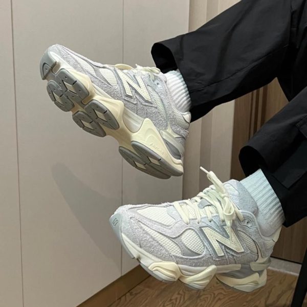 Light gray New Balance sneakers with a chunky off-white sole, worn with white socks, indoors on a wooden floor