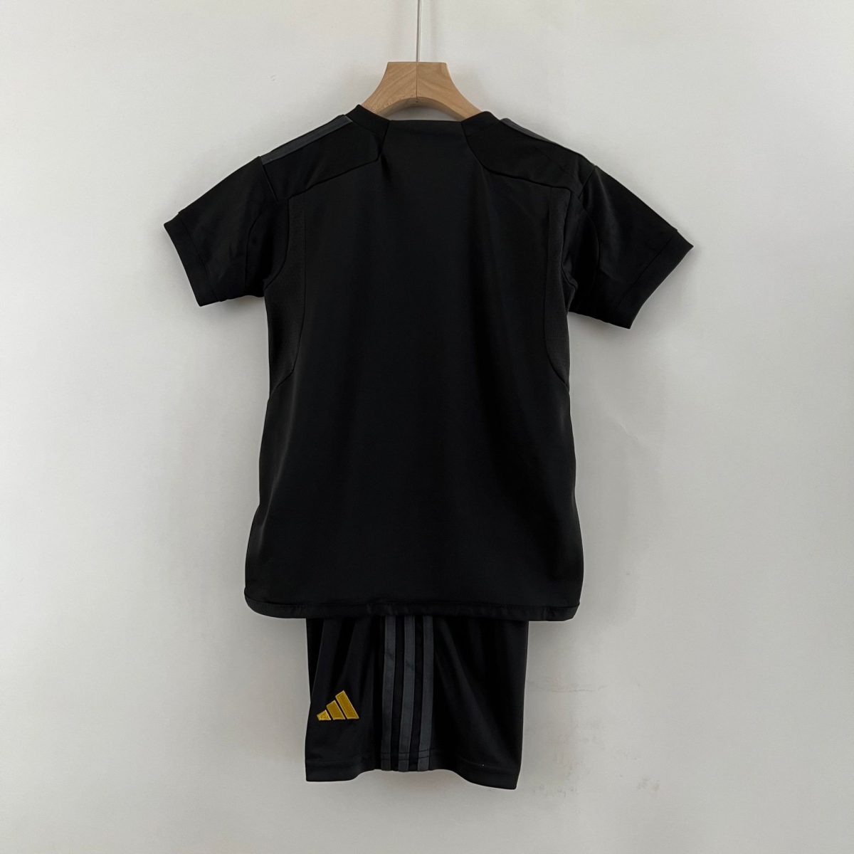 Back view of a black garment on a wooden hanger against a pale wall; a pair of black pants with a small yellow Adidas logo visible at the bottom edge.