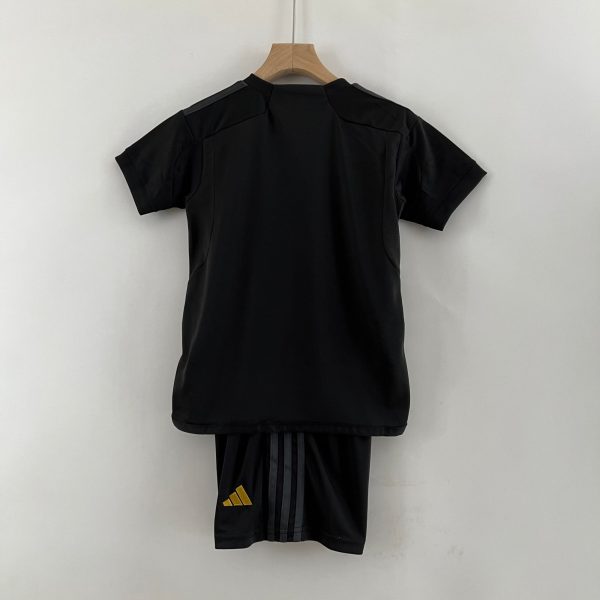 Back view of a black garment on a wooden hanger against a pale wall; a pair of black pants with a small yellow Adidas logo visible at the bottom edge.