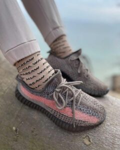 Person wearing gray knit sneakers with a pink stripe, standing on a concrete surface near a body of water, socked feet visible.