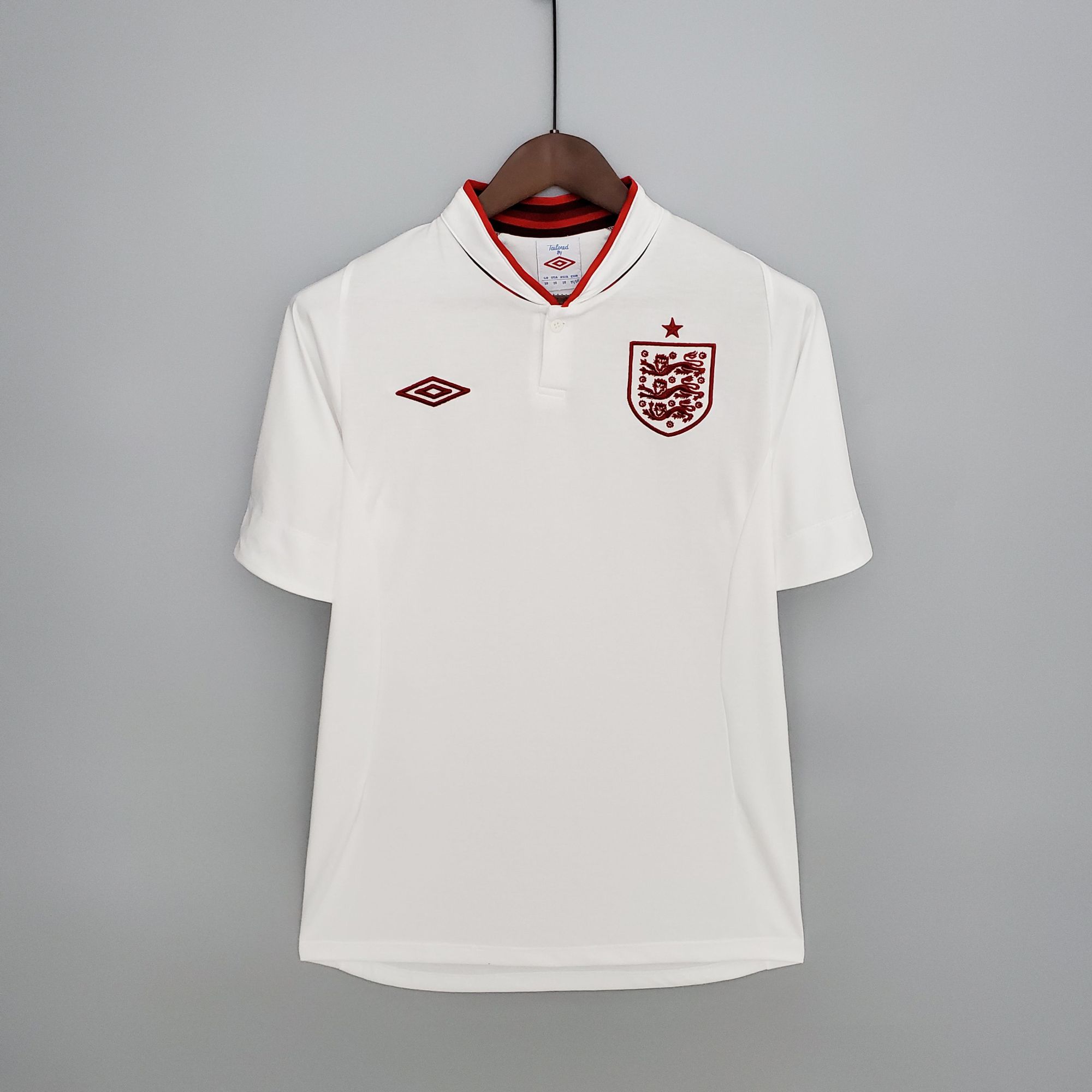 d66e4e62_92df8217-9604-439e-933a-7fd7cf7b5514 | UAE SPORT STORE White England national football team jersey hanging on a wooden hanger against a gray wall, front view