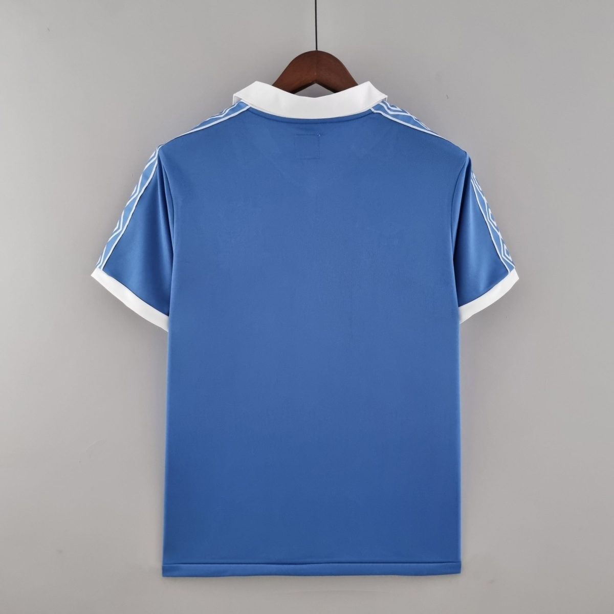 Back view of a blue polo shirt on a wooden hanger, white collar and white cuff trim visible against a gray backdrop.