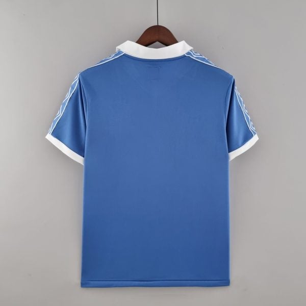 Back view of a blue polo shirt on a wooden hanger, white collar and white cuff trim visible against a gray backdrop.