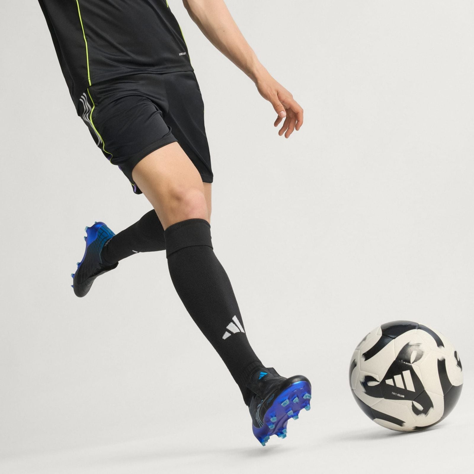 d74cd0f0-a76d-4893-b64e-d5f206466563 | UAE SPORT STORE Soccer player in black kit with neon piping kicking a soccer ball in mid-motion on a white background, blue cleats visible.