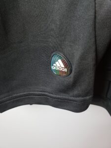 Close-up of black fabric with an oval Adidas logo badge featuring rainbow curved lines and white lettering near the bottom edge of the garment.