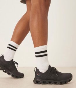 Person wearing black athletic shoes with a chunky, segmented sole and white crew socks with two black stripes walking or posing.