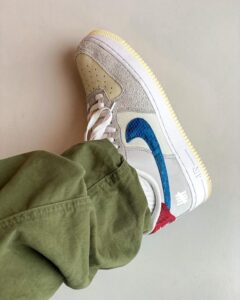 Close-up of a sneaker showing a blue swoosh, beige suede panels, white laces, and a pale gum sole, worn with olive-green pants.