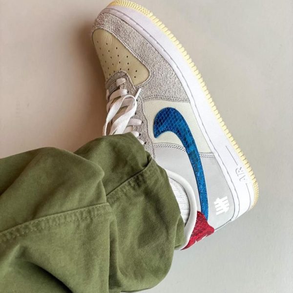 Close-up of a sneaker showing a blue swoosh, beige suede panels, white laces, and a pale gum sole, worn with olive-green pants.