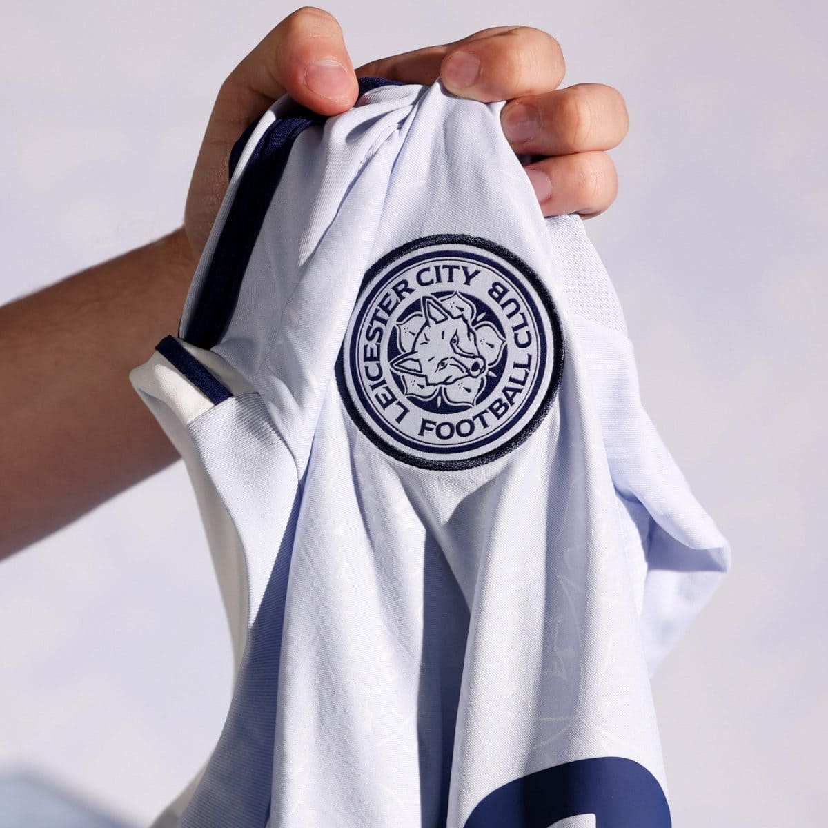 A hand holds up a white Leicester City Football Club jersey, displaying the club crest with a fox head logo on the chest area.