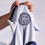 A hand holds up a white Leicester City Football Club jersey, displaying the club crest with a fox head logo on the chest area.