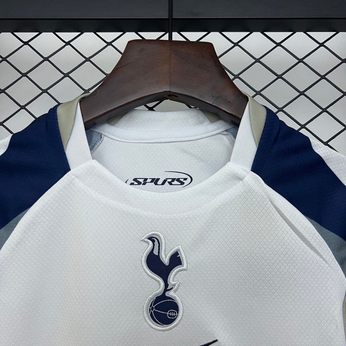 Light gray sports shirt on a dark wooden hanger, navy trim visible, with a 'Spurs' logo on the chest against a chain-link fence background.