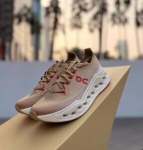 Beige knit running shoes with red laces and a chunky white sole displayed on a beige pedestal.