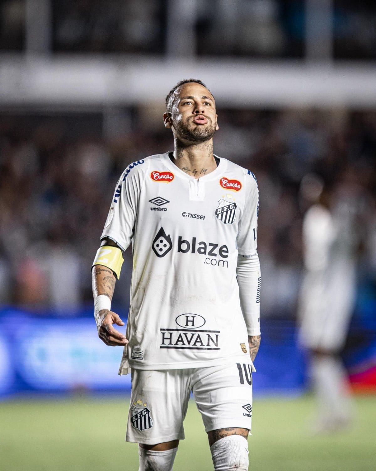 Soccer player in a white Santos FC kit on the field, blowing a kiss with a yellow captain's armband visible and a blurred crowd behind him.