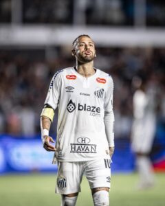 dE5SqOpbKHKc9oW | UAE SPORT STORE Soccer player in a white Santos FC kit on the field, blowing a kiss with a yellow captain's armband visible and a blurred crowd behind him.