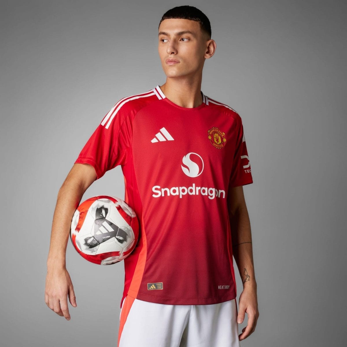 Man wearing a red Manchester United jersey holds a soccer ball under his left arm, posing against a gray backdrop.
