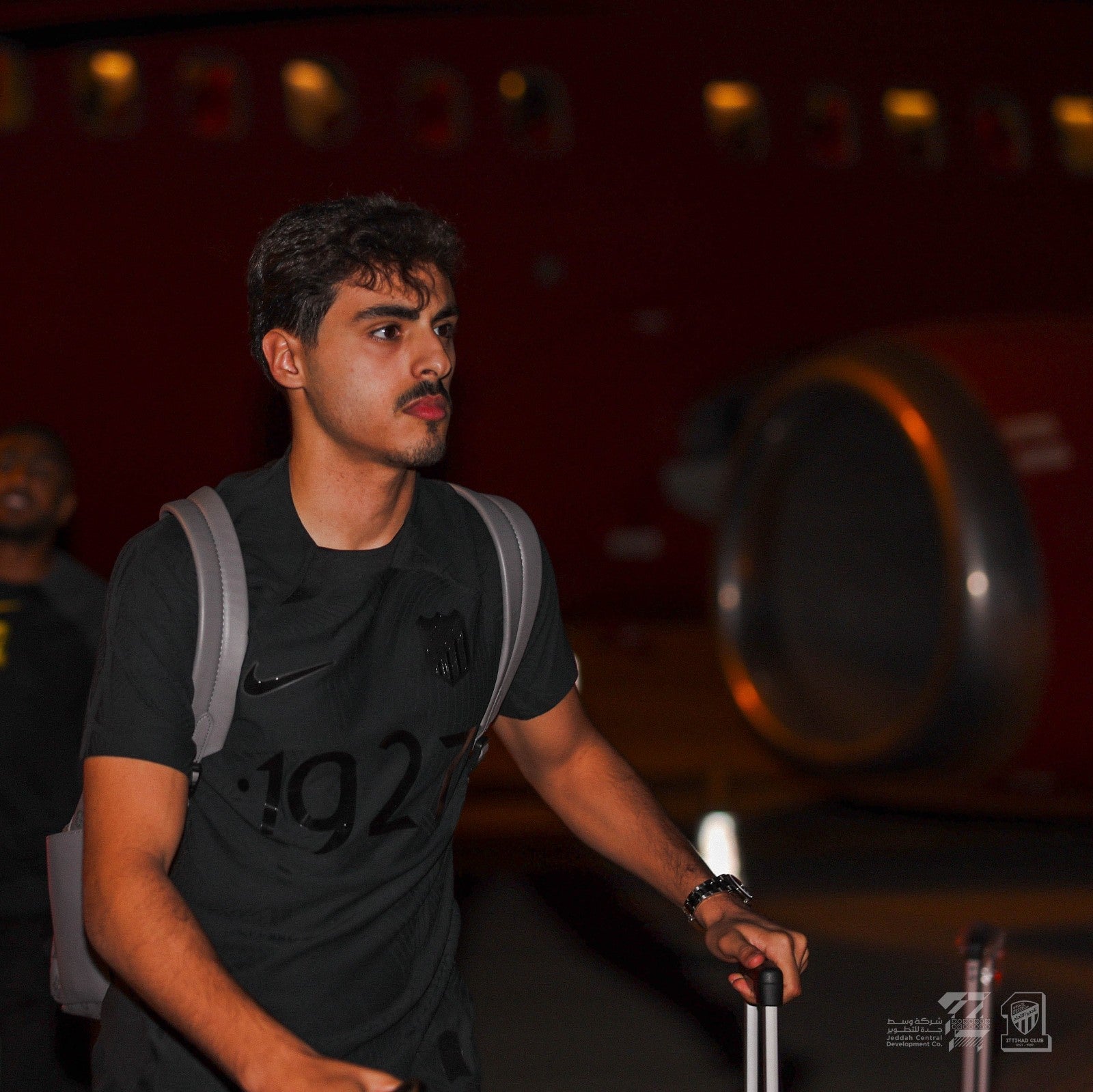 dVWs8gglNYvlEvo | UAE SPORT STORE Young man with a black t-shirt and gray backpack, pushing a rolling suitcase, looking to the side at night.
