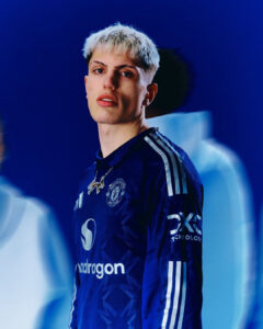 Portrait of a young man with platinum blonde hair wearing a blue Manchester United jersey, looking at the camera.