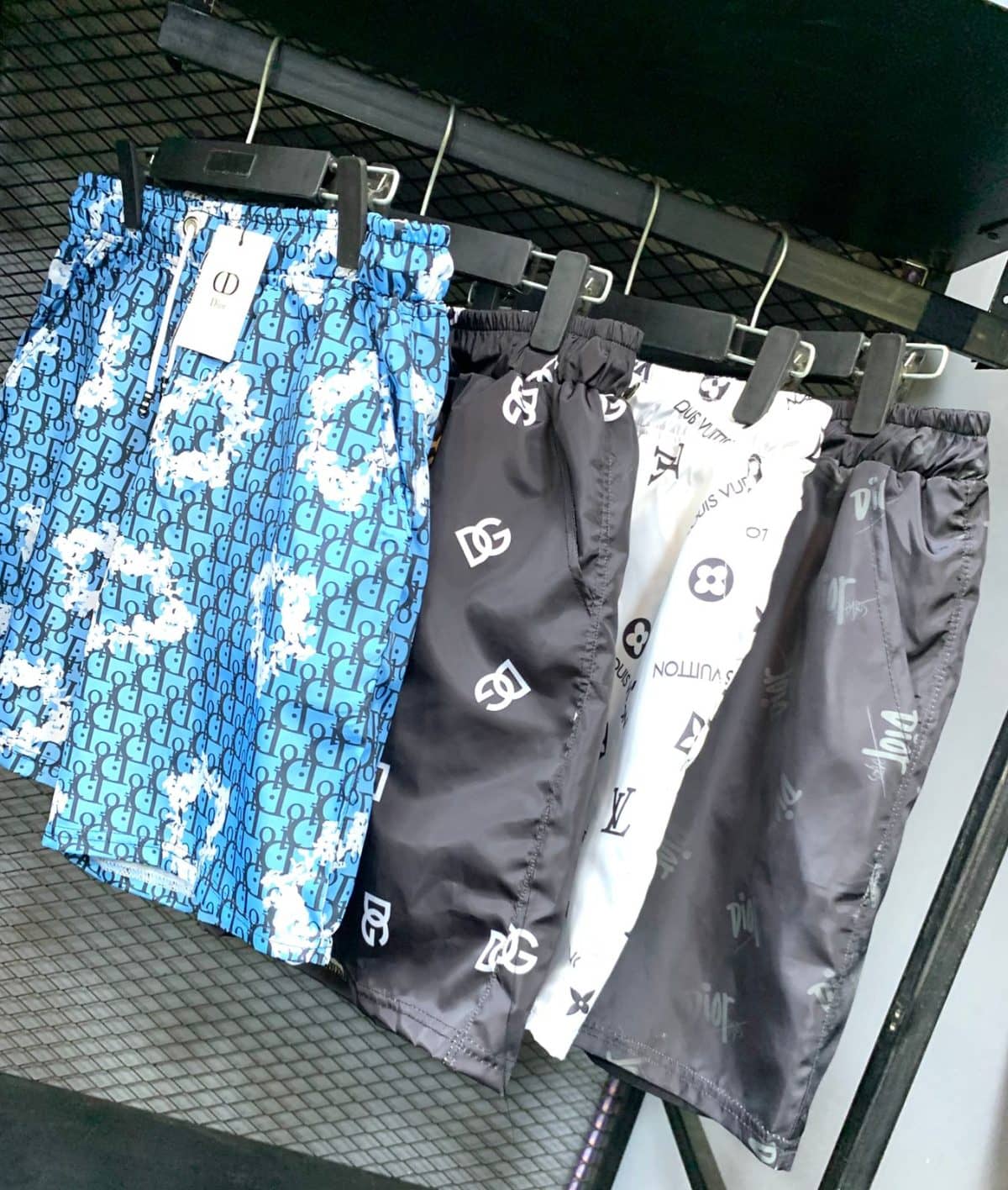 Blue patterned shorts with a Dior tag hanging on a clothing rack in a store window/aisle.
