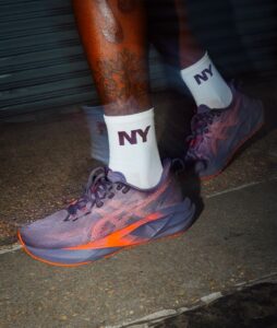 da74bbc1-7370-40b1-92dd-9a5a9c903dba | UAE SPORT STORE Person wearing white NY socks and purple-orange running shoes stepping on pavement; visible tattoos on lower leg.