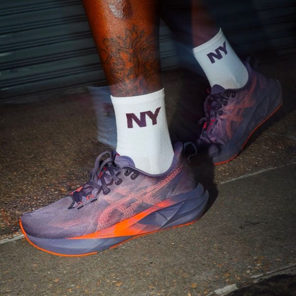 Person wearing white NY socks and purple-orange running shoes stepping on pavement; visible tattoos on lower leg.