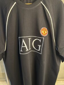 Black athletic shirt with a prominent white 'AIG' logo on the chest and a small Manchester United crest badge above it, white piping along the seams.