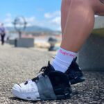 Close-up of a two-tone Nike sneaker on a gravel path, with a white sock showing a red 'Supreme' logo, outdoors by a waterfront.