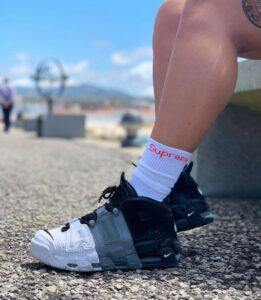 Close-up of a two-tone Nike sneaker on a gravel path, with a white sock showing a red 'Supreme' logo, outdoors by a waterfront.