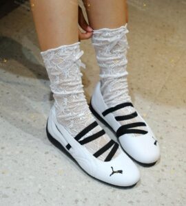 db3fa394-bc52-456b-adaa-74115c762c51 | UAE SPORT STORE Feet wearing white lace knee-high socks with small bows, paired with white strappy flats on a speckled floor.