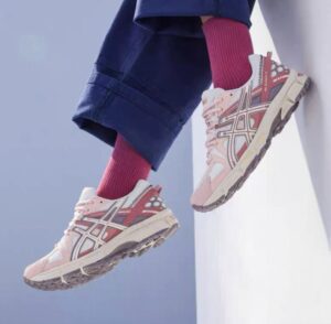 Pink and white athletic sneakers with red detailing, worn with bright pink socks and dark blue pants, mid-air against a light wall.