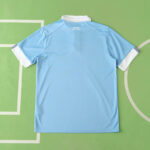 Light blue short-sleeve sports jersey laid flat on a green background, back side facing up, white cuffs and collar visible.