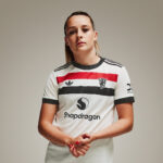 Person wearing a white sports jersey with red and black stripes, standing with hands clasped in front.