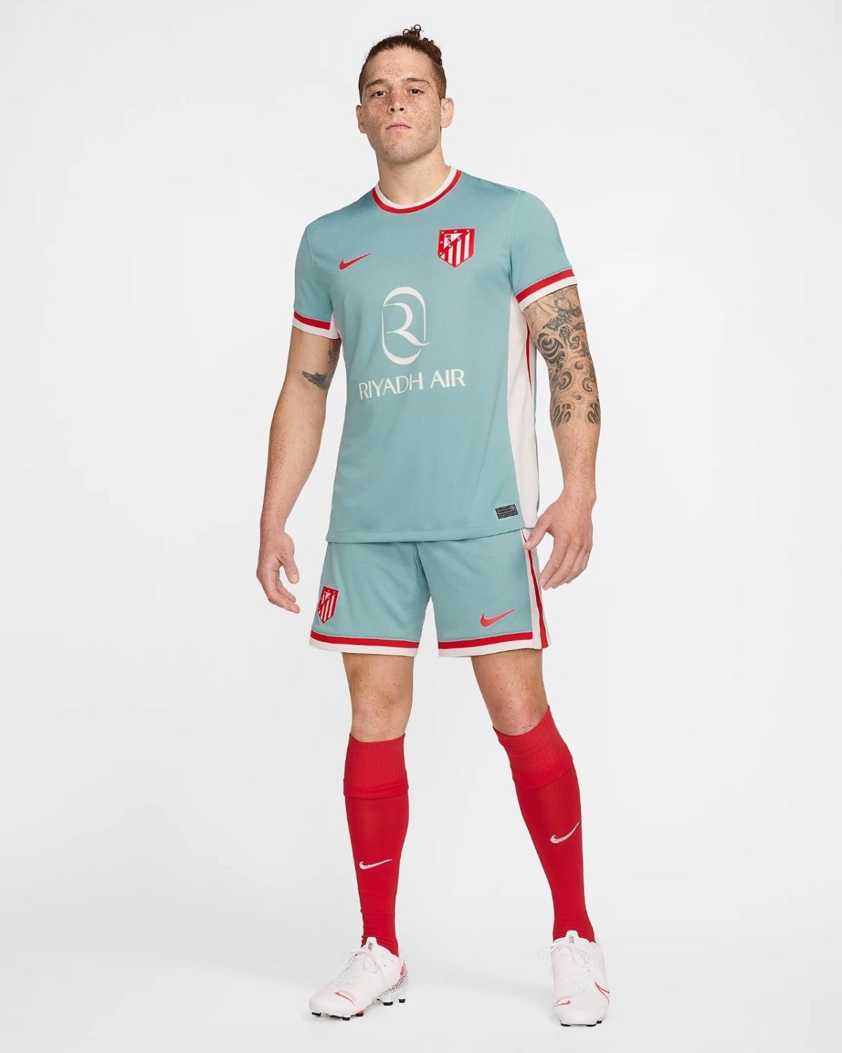Male footballer wearing an Atlético Madrid away kit in light blue with red trim, red socks, and white cleats, posing for a product photo.