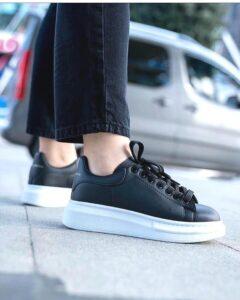 dc82dd22-f1d2-43ca-9e00-071e74087de9_6fc606e9-30fa-4985-b31f-51ef36a934f1 | UAE SPORT STORE Person wearing black leather sneakers with a thick white sole, standing on a sidewalk with a city street in the background.