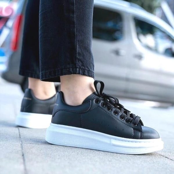 Person wearing black leather sneakers with a thick white sole, standing on a sidewalk with a city street in the background.