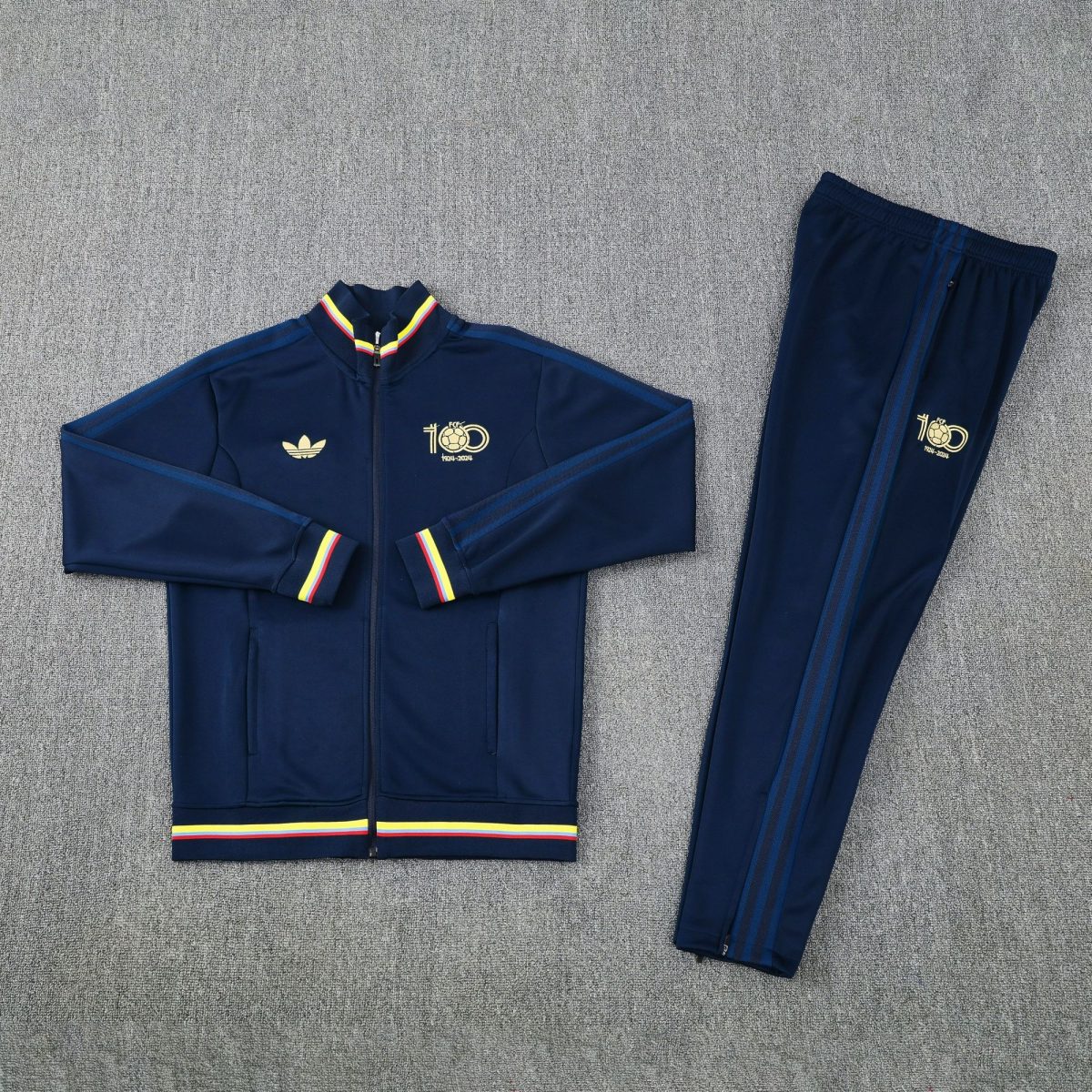 Navy tracksuit jacket and pants laid flat, featuring gold crest and rainbow-yellow-red trim on cuffs and hem, athletic set.