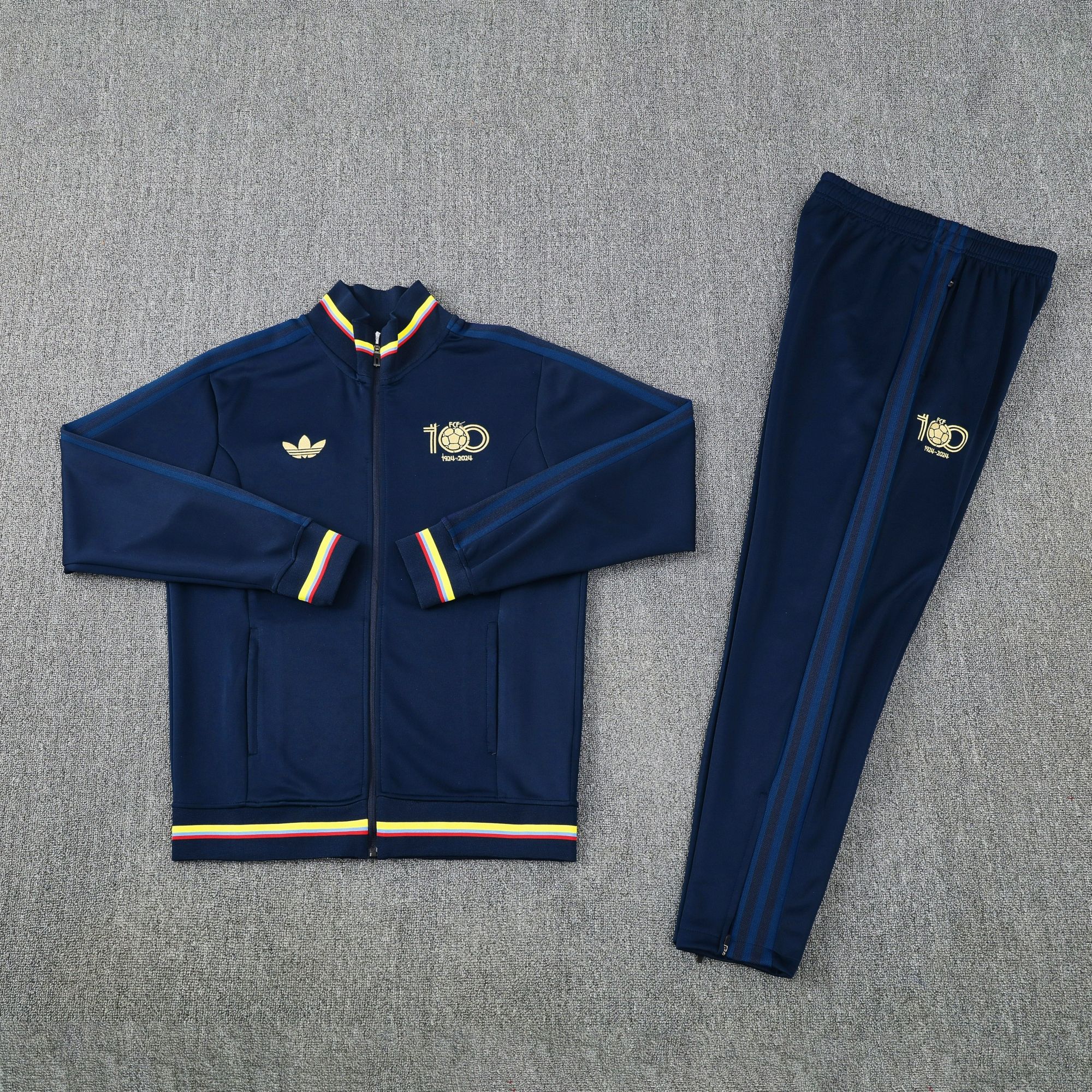 dca709f5 | UAE SPORT STORE Navy tracksuit jacket and pants laid flat, featuring gold crest and rainbow-yellow-red trim on cuffs and hem, athletic set.