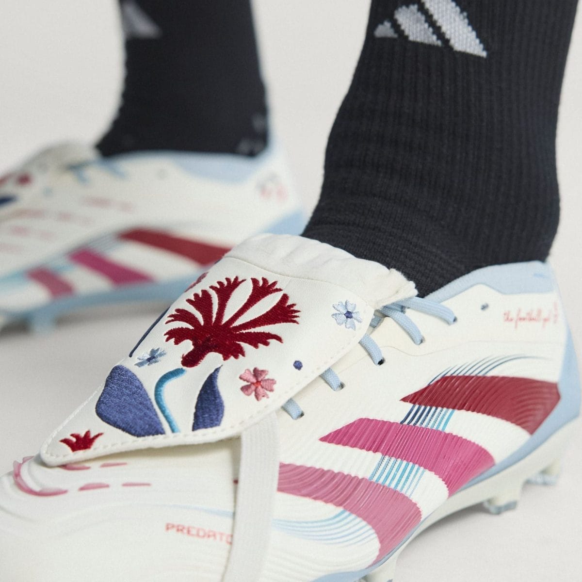 Close-up of a white sneaker with pink and blue stripes, light blue laces, and red coral and floral embroidery worn with a black sock beneath.
