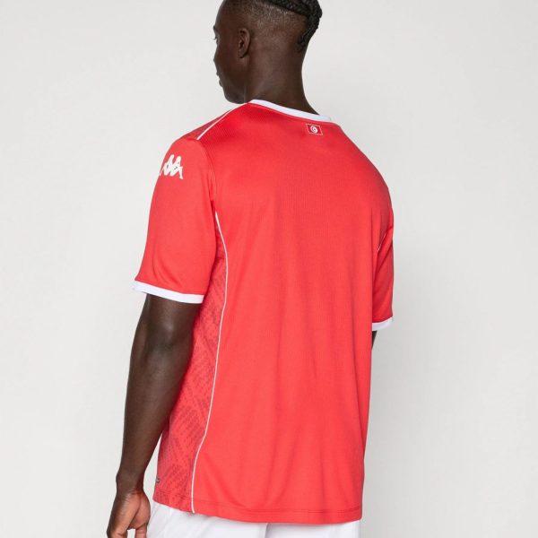 Back view of a dark-skinned man wearing a red athletic jersey with white trim and white shorts, standing against a plain light background, showing braided hair.