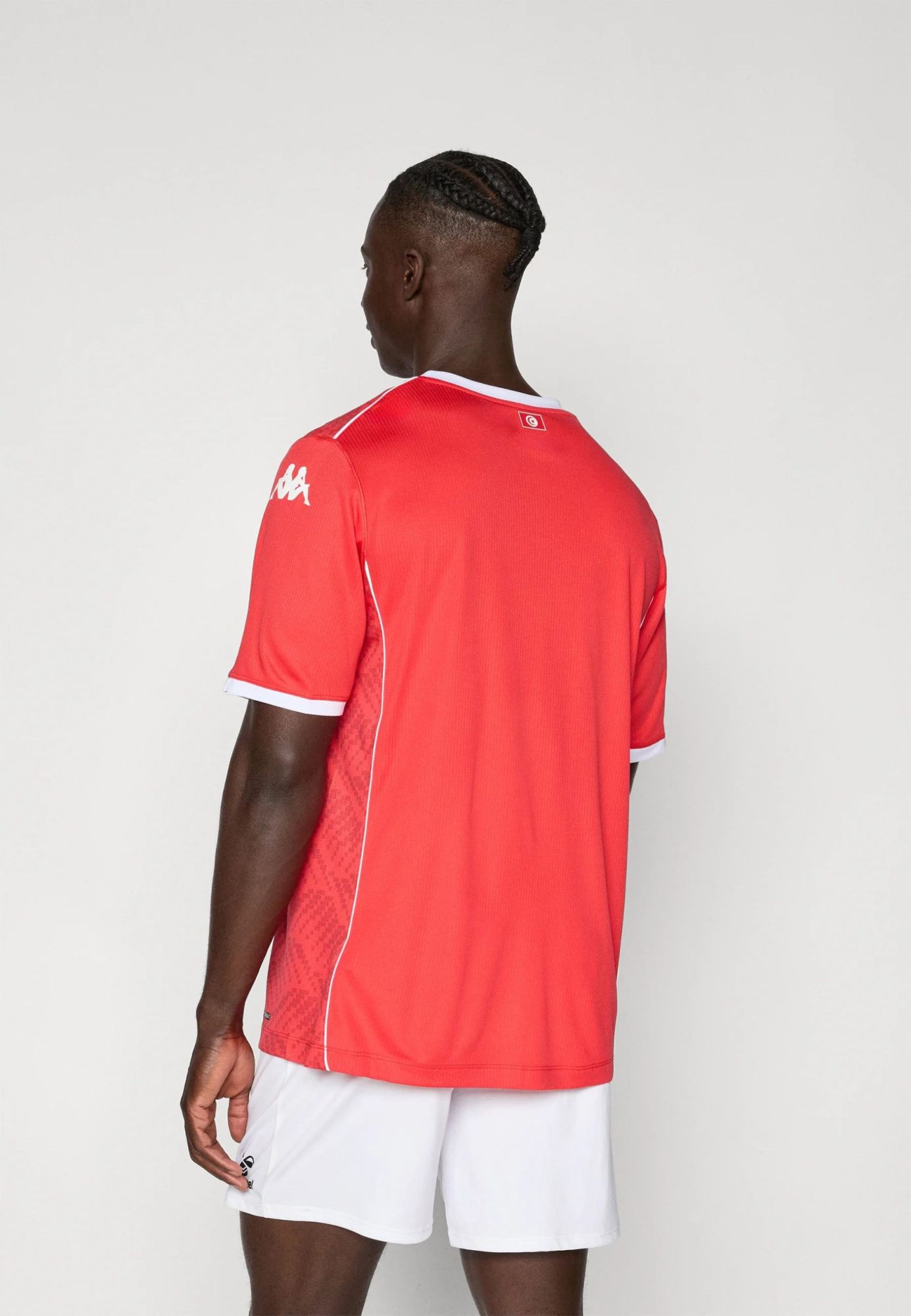 de0eed70235745d6949fc585e0657d63 | UAE SPORT STORE Back view of a dark-skinned man wearing a red athletic jersey with white trim and white shorts, standing against a plain light background, showing braided hair.