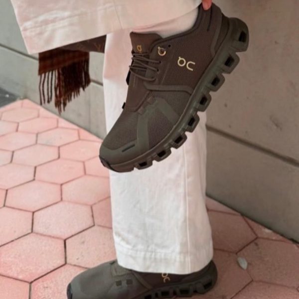 Olive green athletic sneaker with a chunky segmented sole, worn with white pants as the person lifts their foot.