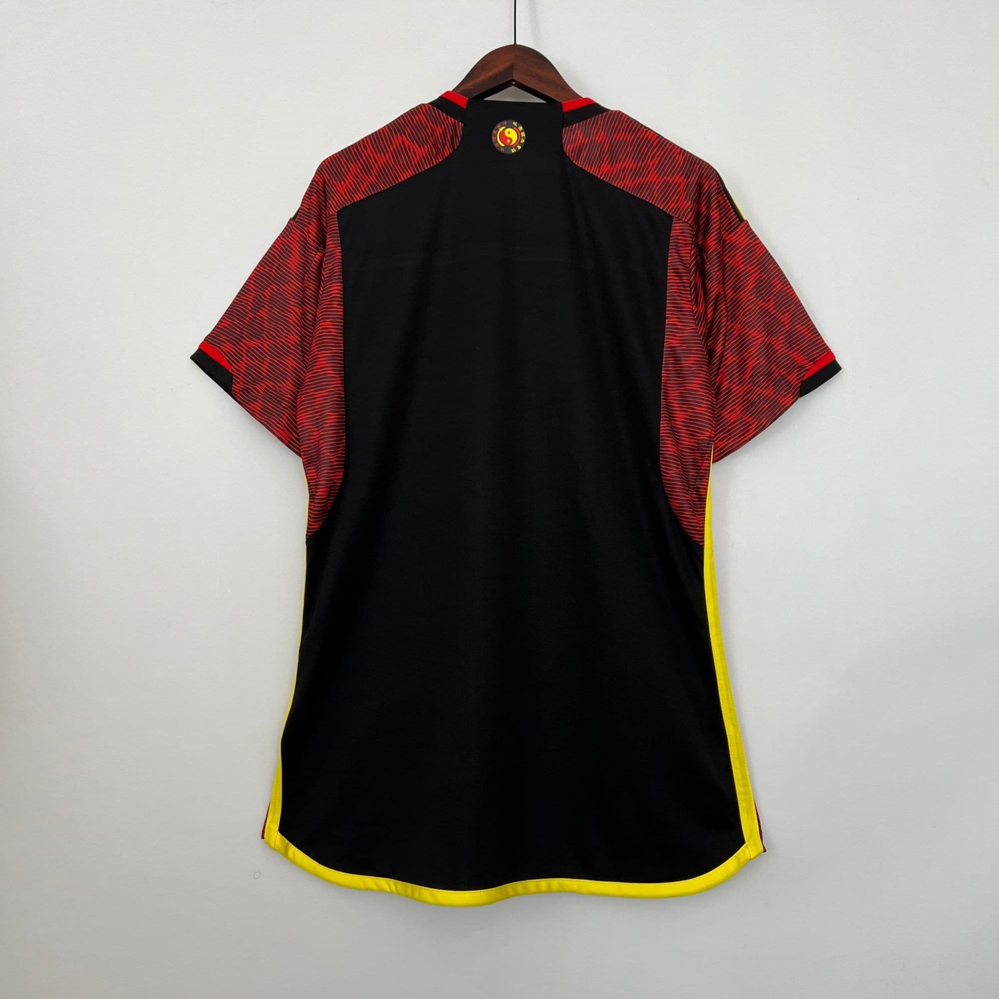 de937b0e_17756da3-1746-4d97-9121-99d1e3f20aaf | UAE SPORT STORE Back view of a black sports jersey with red patterned sleeves, bright yellow trim along the hem, hanging on a wooden hanger, logo at the neck.